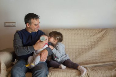 Evdeki kanepede oturan bir babanın yeni doğmuş oğlunu kucağında tutarken Bineron ve ablasının ona sarılıp öptüğü görüntüsü. Kardeş sevgisi ve sevgi dolu bir aile.