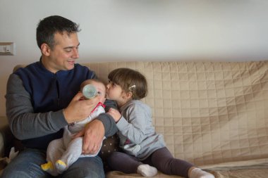 Evdeki kanepede oturan bir babanın yeni doğmuş oğlunu kucağında tutarken Bineron ve ablasının ona sarılıp öptüğü görüntüsü. Kardeş sevgisi ve sevgi dolu bir aile.