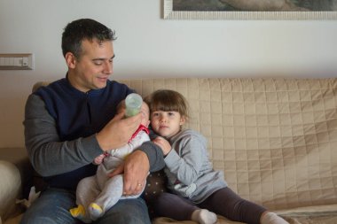 Evdeki kanepede oturan bir babanın yeni doğmuş oğlunu kucağında tutarken Bineron ve ablasının ona sarılıp öptüğü görüntüsü. Kardeş sevgisi ve sevgi dolu bir aile.