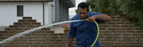Image of a man in his home garden drinking water from a lawn hose. Reference to waste of water. Horizontal banner
