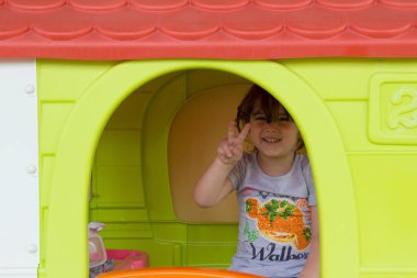 Evinin bahçesindeki bir oyun evinde oynarken parmaklarını zafere gösteren sevimli, gülümseyen küçük bir kızın görüntüsü. Çocuklar için eğlence ve oyunlar