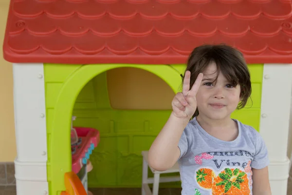 Image of an adorable smiling little girl pointing her fingers to victory while playing in a playhouse in her home garden. Leisure and games for children