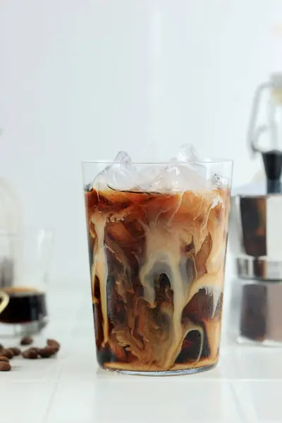 Milk Swirling in a Glass of Black Coffee with Rock Ice on White Table