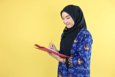 Young ASN Government Worker Wearing Korpri Batik Uniform Holding Pencil, Checking Document Paper. Isolated on Yellow Background  clipart