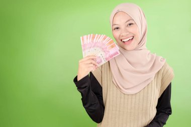Kahverengi kazaklı, Endonezya Rupia Banknotlarıyla nakit para tutan, Gülümseyen ve Kameraya bakan Asyalı Güzel Müslüman Kadın 