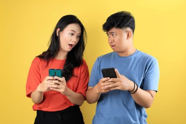 Cep telefonu tutarken ayakta duran genç Asyalı çift şok olmuş. Sarı Arkaplanda İzole Edildi 