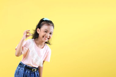 Asyalı Çocuk gülümserken, OK Finger işareti veriyor, Mutlu Neşeli Kız pembe gömlek ve kot pantolon giyiyor, gülüyor ve iyi bir jest yapıyor. Sarı Arkaplanda İzole Edildi