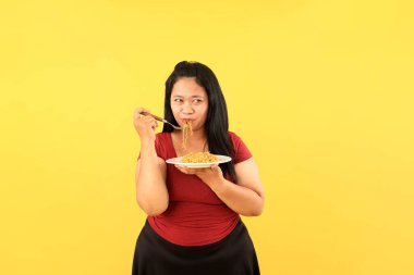 Büyük Şişman Asyalı Kadın Bombalı Göz 'le Anında Erişte yiyor, Sarı Arka plan' da izole edilmiş.