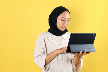 Tebessüm eden, tesettürlü, genç, Asyalı Müslüman bir kadının portresi Dijital Tablet tutan, Tab 'e bakan. Sarı Stüdyo Arkaplanda İzole Edildi