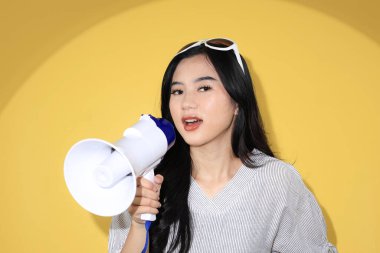 Megafonlu, çizgili beyaz gömlekli ve güneş gözlüklü genç bir kadın, Sarı Stüdyo Arkaplanı ile ilgili bir duyuru yapıyor.