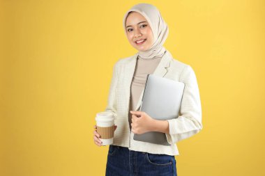 Krema Takımlı Genç Asyalı Başörtülü Kadın ve Dizüstü bilgisayar ve Sıcak Kahve Kupasını Tutan Başörtüsü. Sarı Arkaplanda İzole Edildi