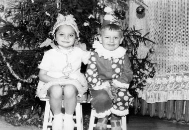 Furstenberg, East Germany - December 31, 1970: Vintage portrait of a little girl and boy in New Year's costumes near a Christmas tree. Retro photography.