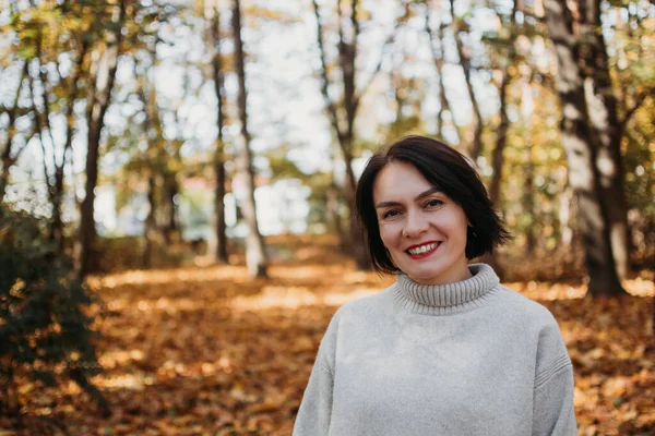 Güneşli bir sonbahar gününde parkta gülümseyen mutlu bir kadın. Güzel bir sonbahar gününde gri kazak giyen neşeli, güzel bir kadın. Doğal ışık. Yüksek kalite fotoğraf.