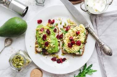 Kahvaltıda sağlıklı avokado tostları ya da öğle yemeğinde çavdar ekmeği, krem peynir, roka, dilimlenmiş avokado, kızılcık, balkabağı, kenevir ve susam tohumu. Vejetaryen sandviçler. Temiz yemek. Üst görünüm.