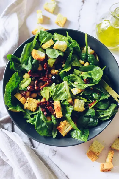 Healthy salad with spinach, croutons, sun-dried tomatoes and black olives in a bowl on white marble background. American style salad. Vegetarian tasty food recipe. Cook book recipe.