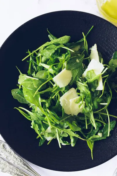 Healthy green salad with fresh arugula, parmesan cheese and olive oil in a dark bowl on marble background. Vegetarian tasty salad recipe. Diet fitness food menu. Recipe for cook book. Top view.