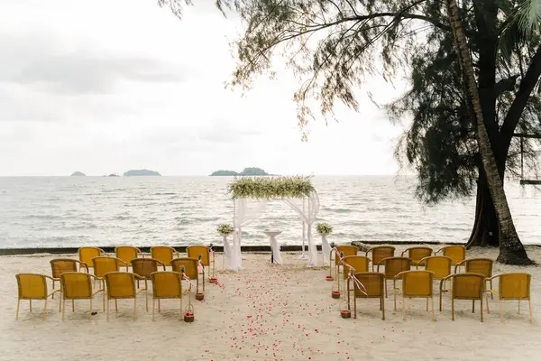Beyaz kumaştan düğün kemeri sandalyeleri ve arka planda deniz bulunan yeşil ve beyaz buketlerle akşam plajı düğün töreni. Düğün töreni, plaj düğün yeri.