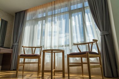 Lüks odada ahşap sandalyeler ve gökyüzü manzaralı beyaz perdelerden oluşan bir arka plan bulunuyor. Sandalye ve perdeli bir otel odasının içi. İçeride kimse yok.