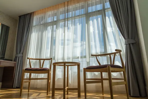 Lüks odada ahşap sandalyeler ve gökyüzü manzaralı beyaz perdelerden oluşan bir arka plan bulunuyor. Sandalye ve perdeli bir otel odasının içi. İçeride kimse yok.