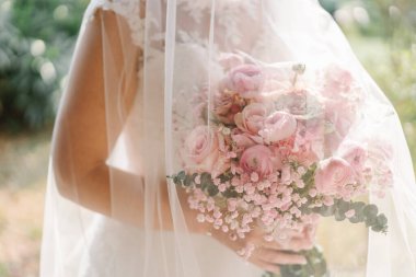 Pembe buketli gelin beyaz peçeli beyaz bir elbise giyiyor. Buket pembe çiçeklerden, muhtemelen güllerden ya da şakayıklardan ve daha küçük beyaz çiçeklerden oluşur, muhtemelen gypthe (bebek nefesi).).