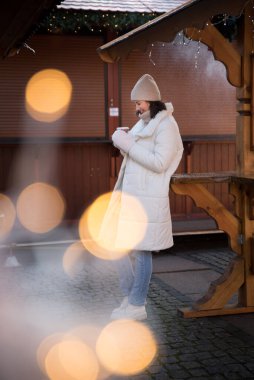 Adil. Beyaz ceketli ve beyaz şapkalı bir kız. Kız gülümsüyor. Elde bir fincan kahve. Ahşap yazlık. Kız seviniyor. duygular.