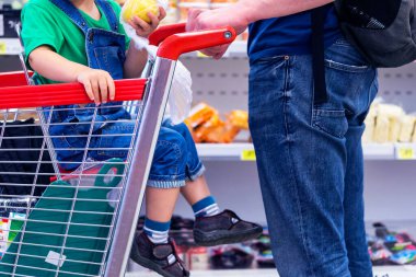 Çocuk dükkanda tekerlekli sepette oturuyor. Süpermarkette çocuklu bir adam. Baba ve oğlu rafların arkasında ürünlerle birlikte. Konsept alıcılar. Müşteriler, alışveriş konsepti
