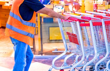 Adam bir süpermarkette bir yığın el arabası itiyor. Alışveriş merkezinin çalışanı market arabalarını taşıyor. Bir sürü alışveriş arabası. Müşteriler, müşteriler