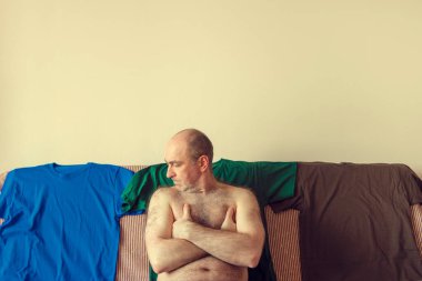  Çıplak adam kanepede oturuyor. Bir adam ve birkaç tişört. Kıyafet seçimi ve uygun kıyafetler.