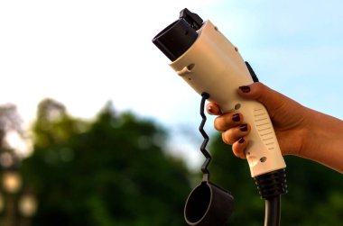 Bir kadın eli, elektrikli bir arabanın kablosunu gökyüzü ve doğanın arka planına karşı tutar. Sürücü elektrikli bir aracı şarj etmek için bir kablo tutuyor. Temiz alternatif enerji kavramı
