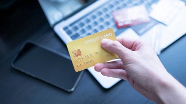 A hand holds a credit card in front of a laptop, suggesting online shopping or banking. cards and a smartphone on the desk, emphasizing the concept of digital payments and financial transactions. clipart