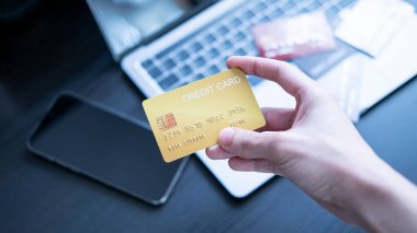 Bir el dizüstü bilgisayarın önünde kredi kartı tutuyor, online alışveriş ya da bankacılık olduğunu gösteriyor. Kartlar ve masanın üzerinde dijital ödemeler ve finansal işlemler kavramını vurgulayan bir akıllı telefon.