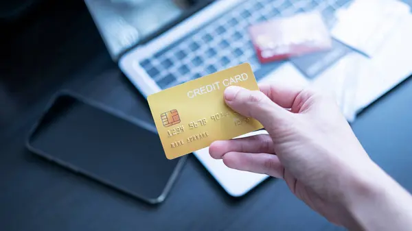 Bir el dizüstü bilgisayarın önünde kredi kartı tutuyor, online alışveriş ya da bankacılık olduğunu gösteriyor. Kartlar ve masanın üzerinde dijital ödemeler ve finansal işlemler kavramını vurgulayan bir akıllı telefon.