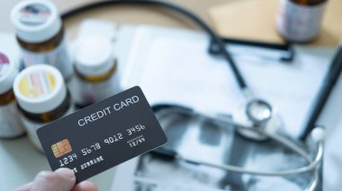 Steteskop ve ilaç şişelerinin yanında siyah kredi kartıyla yakın plan çekilmiş. Hastane faturası..