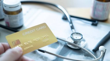 Tıbbi belgeler ve steteskop yanında altın kredi kartı tutan bir el, ödeme seçeneği.