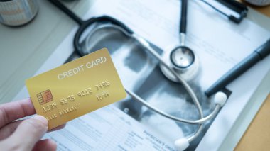 Steteskopun üstünde siyah bir kredi kartı ve tıbbi kayıtları tutan bir el..