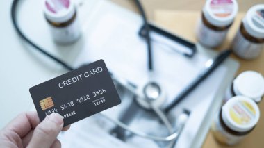 Tıbbi faturalar ve ilaç şişelerinin üzerinde, sağlık ödemelerini ve mali baskıyı simgeleyen siyah bir kredi kartı bulunduruyor.