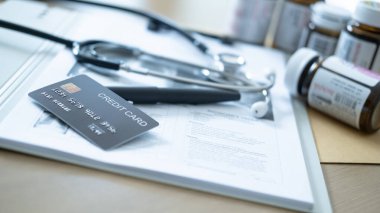 Gri bir kredi kartının üstüne haplar ve steteskopla yakın mesafeden çekilmiş gri bir fotoğraf..