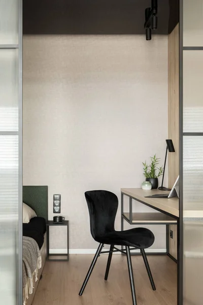Elegant black chair in home office room with wooden desk, bed and reinforced glass door