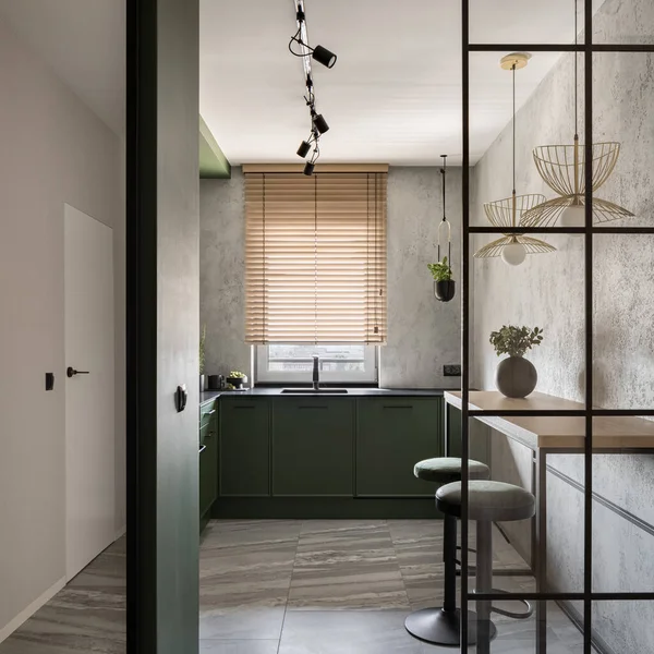 Modern kitchen with stylish green furniture, gray walls, window with wooden blinds and modern reinforced glass wall