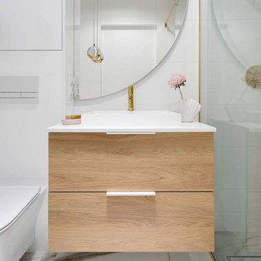 Close-up on wooden drawers under bathroom washbasin with golden tap, decorations and big semicircle mirror