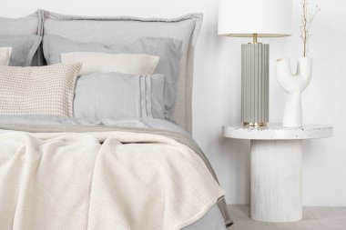 Stylish lamp and decorative vase on white bedside table next to cozy bed with elegant bedclothes