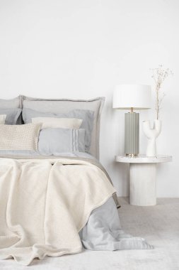 Elegant lamp and decorative vase on bedside table next to comfortable bed with flax bedclothes in bright bedroom
