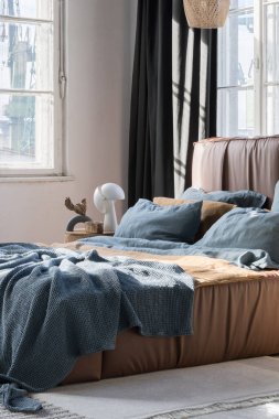 Sunny bedroom with big bed in brown leather frame with blue and brown bedclothes and many stylish decorations