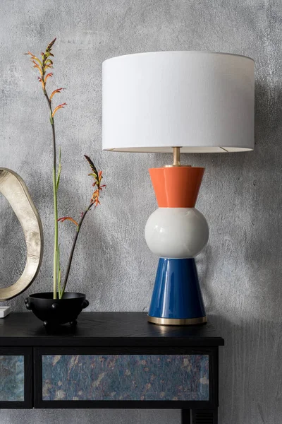 Elegant and decorative lamp in white, orange and navy blue next to stylish decorations on black console table