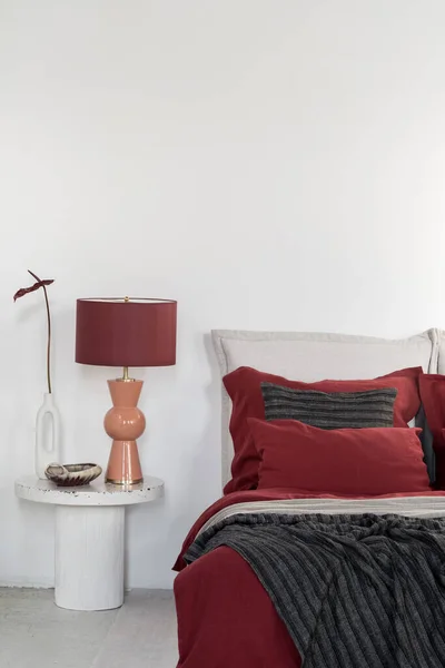 精致的橙色和红色的灯和花瓶 在大理石床头柜上绽放着花朵 旁边是舒适的床 明亮的卧室里有红黑相间的床上用品和枕头 — 图库照片