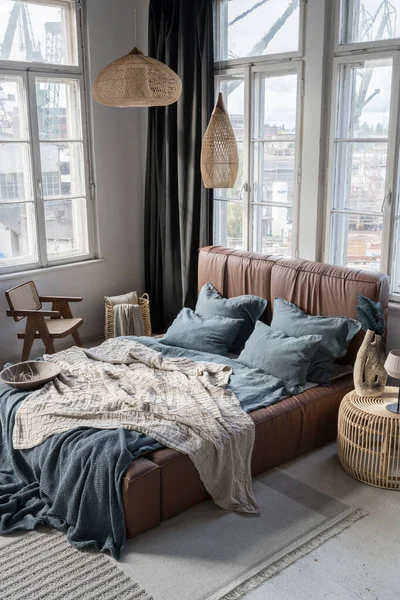 Spacious and eclectic bedroom with big windows, cozy bed with brown, leather headboard and blue, flax pillows and bedclothes and rattan lamps, chair and bedside table