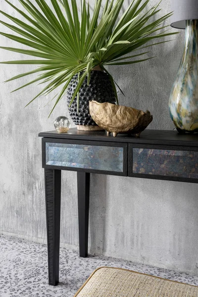 Elegant, black console table with drawers, aesthetic decorations, golden bowl, vase with green leaves and lamp in room with gray wall and patterned carpet