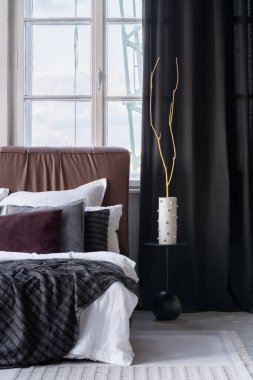 Siyah komodinin üzerinde sarı dalları olan modern bir vazo. Yatak odasında büyük pencereli deri yatak başlığı olan rahat yatağın yanında.
