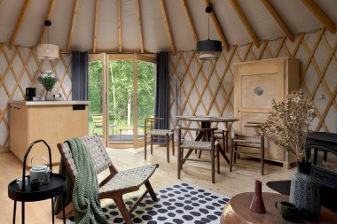 Beautiful interior of yurt tent with kitchen area, dining table with chairs and balcony doors open to forest clipart