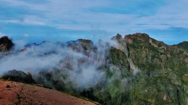 Yeşil bitkilerle kaplı görkemli kayalık dağlar, açık mavi bir gökyüzünün altında sürüklenen sisle kaplanmış. Yükseliş, doku ve sakin atmosferle dolu sakin ve sinematik bir manzara..